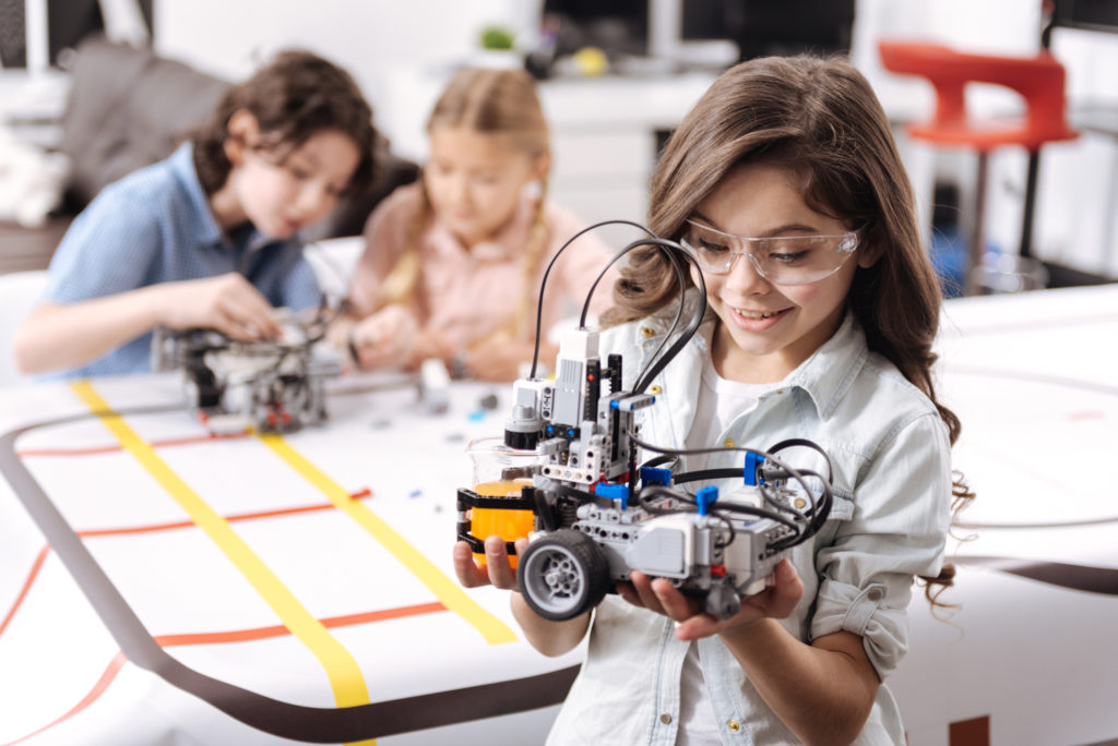Smart generation exploring science. Sincere smiling emotional girl standing at school and holding electronic robot while her colleagues working on the project