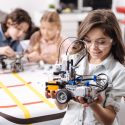 Smart generation exploring science. Sincere smiling emotional girl standing at school and holding electronic robot while her colleagues working on the project